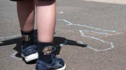 A student's legs on a sunny playground. In front of them, their shadow is traced on the ground in blue chalk. 