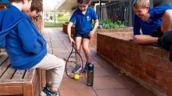 3 seated students observe as a 4th hits a ball on the ground with a racket 
